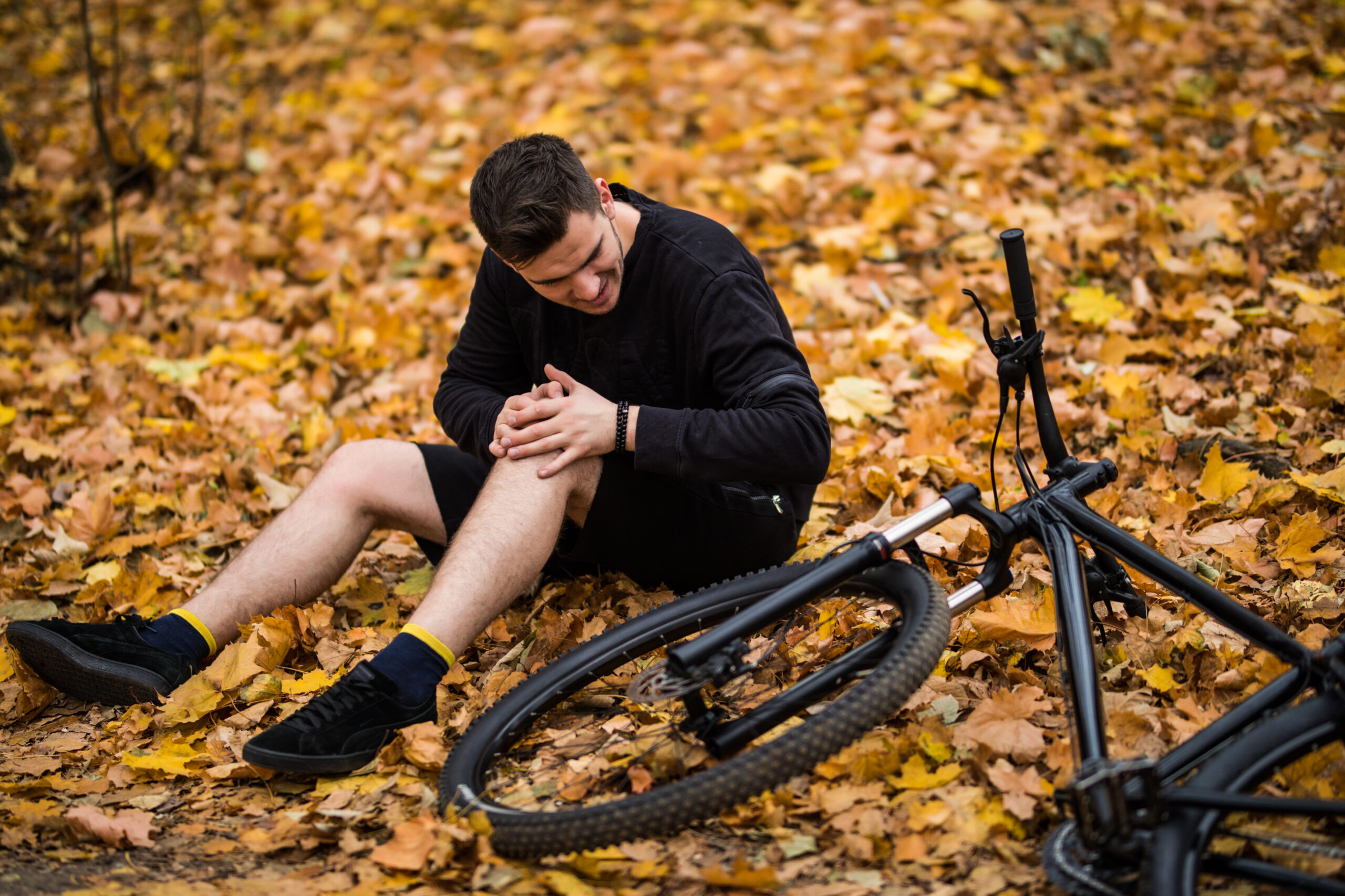 Active young man holding by his hurt or broken leg while lying on forest path by his bicycle